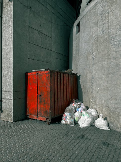 A red metal waste skip with rusted surfaces and vertical ridges is positioned against a corner of a dark gray facade with large, smooth stone or concrete panels and a lighter gray brick wall. The skip is placed on a textured, dark rubber or stone pavement, and several clear plastic bags filled with mixed rubbish are jumbled beside it, spilling slightly onto the ground. The environment appears to be an urban exterior setting, possibly an alleyway or service area behind a building, with minimal signage or other objects visible. The lighting is natural, diffusing evenly across the surfaces, highlighting the weathered condition of the skip and the assorted waste bags, which are typical of private or independent waste collection methods. This scene visually illustrates private rubbish disposal or on-site clearance, exemplifying alternative waste handling beyond municipal collection services, consistent with professional waste management practices in urban areas, especially in affluent neighborhoods like Knightsbridge.