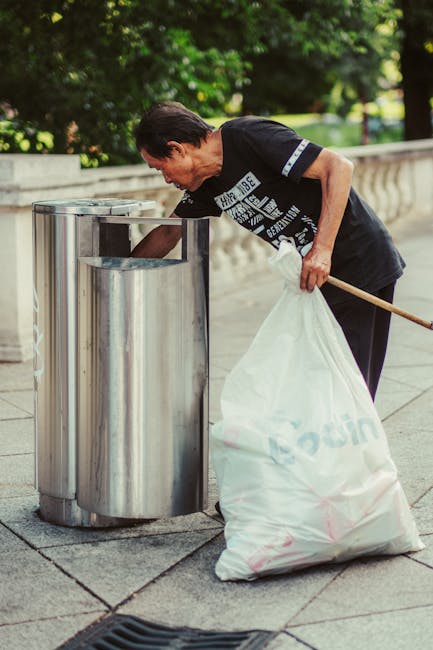 A middle-aged man with dark, slightly long hair, dressed in a black T-shirt with a white graphic print on the front, is standing outdoors on a paved pathway near a stone balustrade. He is leaning forward, using his right arm to reach into a metallic recycling bin with a rounded, cylindrical shape divided into separate compartments, likely for different types of waste. In his left hand, he holds a large white plastic bag filled with various rubbish, and a wooden stick or broom handle is tucked under his arm. The surrounding environment includes lush green trees and foliage, providing a natural backdrop, and sunlight filters through the leaves, casting soft light on the scene. The area appears to be a public park or walkway, emphasizing an act of independent waste management, typical of private rubbish clearance services or on-site rubbish collection efforts, aligned with professional waste removal practices in urban outdoor settings.