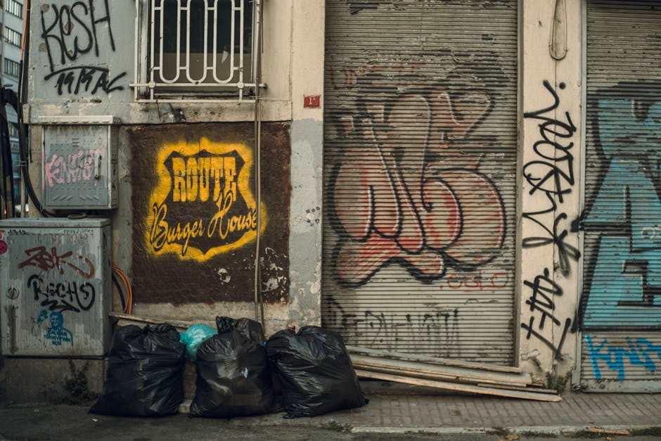 A narrow urban alleyway cluttered with large black and beige waste bags piled against a makeshift barrier of cardboard boxes and plastic sheeting at the rear, indicating a private rubbish collection or on-site clearance. The ground is uneven asphalt with visible dirt, stains, and small debris scattered throughout. Tall brick and metal-clad walls flank the alley, with some graffiti on the right side. The left wall features weathered bricks, while the right environment appears more industrial, with utility meters and metal railings. Bare tree branches extend overhead, suggesting a late autumn or winter setting. Natural light illuminates the scene from above, casting subtle shadows. This scene visually supports the practice of independent rubbish removal services, such as those offered by companies like rubbishcollectionknightsbridge.org.uk, as part of alternative waste handling outside of standard municipal disposal methods.