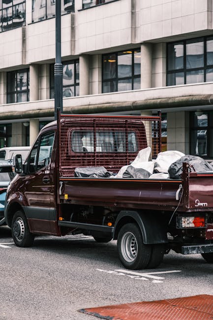 A dark red flatbed utility truck parked on a city street in front of a modern building with large glass windows. The truck's cargo area is filled with various pieces of waste materials, including large black and white garbage bags and miscellaneous debris. The truck features a metal cage at the back, which is partially visible, and has a compact, utilitarian design suitable for rubbish collection or on-site waste removal. The vehicle is positioned close to the curb, with a white car parked slightly behind it. The surrounding environment includes a lamppost and grey pavement, indicating an urban setting. The scene suggests a private waste collection or rubbish removal service, possibly organized by an independent provider offering alternative disposal options beyond standard municipally authorized collection, as implied by the context of alternative waste handling services.