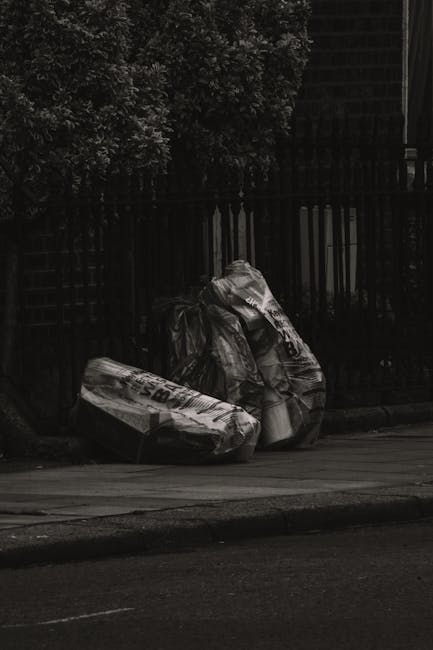 A black-and-white image showing two large black plastic rubbish bags placed on the pavement beside a black metal fence. The bags appear to be filled with waste materials and are leaning slightly, with one bag partially resting on the other. The fencing behind them is composed of vertical metal bars, and there is a dense, leafy tree visible in the upper left corner of the scene. The pavement surface is smooth, and a section of the road is visible in the foreground, indicating an outdoor urban or residential setting. The lighting suggests daytime, with natural ambient light casting minimal shadows. The image subtly relates to waste collection and rubbish disposal services, such as those provided by independent rubbish removal companies like [COMPANY_NAME], which operate outside standard local authority collection systems.