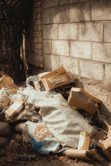 A pile of assorted rubbish, including cardboard boxes, paper bags, and plastic packaging, is placed on the ground next to a concrete block wall and a tree trunk. The cardboard boxes vary in size and are partially open, revealing some contents inside. The paper bags and packaging are crumpled and loosely stacked, with some resting on a textured, off-white fabric bag. The background features a light gray, weathered concrete block wall with visible mortar joints, and a section of dark tree bark is visible on the left side of the image. The scene appears to be an outdoor area adjacent to a building or wall, possibly part of a private property, where waste has been temporarily gathered for collection or disposal. The ground is uneven, with loose soil and small stones, and the lighting is natural, indicating daytime. This scene exemplifies a typical collection of waste material awaiting removal, aligned with private rubbish disposal services like those offered by Rubbish Collection Knightsbridge, which are alternatives to local authority rubbish collection methods.