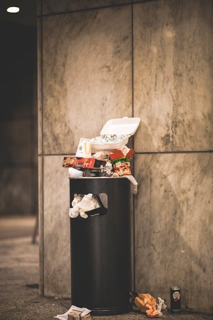 A black cylindrical rubbish bin with a partially open white foam food container resting on top, surrounded by various food packaging and waste items, situated against a large beige tiled wall in an indoor or underground environment. There are crumpled paper and food wrappers spilling onto the floor, along with a discarded drink can and a crumpled snack packet nearby. The scene suggests improper disposal of food waste and packaging in a public area, which could indicate a need for more effective rubbish collection or on-site clearance services; the image reflects challenges in maintaining cleanliness and proper waste handling in busy urban spaces, such as those served by independent rubbish removal providers like [COMPANY_NAME]. The environment appears dimly lit, with the focus on the litter, emphasizing the importance of regular rubbish removal in private or communal spaces to support better waste management practices.