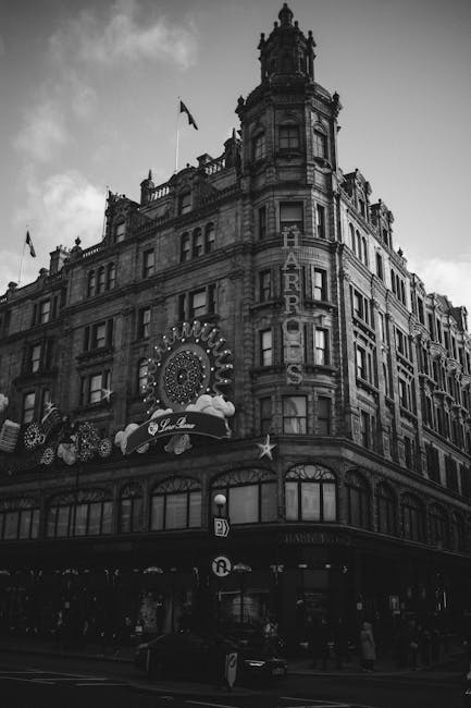 A black and white photograph of a historic urban building with ornate architectural details, including decorative cornices, tall windows, and a rounded corner tower topped with a spire. The building features illuminated signage for a hotel, with the word 'Harrods' vertically displayed on a marquee-style sign along the rounded corner, and a large decorative circular sign with lights above the entrance. The ground floor appears to be a commercial space, with storefront windows and awnings. The scene is set on a city street with parked cars and pedestrians visible at the pavement level. The overall atmosphere is based on the building's classic design and the lively street environment, providing context for how private waste collection and rubbish removal services might be necessary for maintaining such historic urban sites, especially when managing trash during renovations or street events. The muted tones emphasize the architectural features and urban setting, aligning with the context of alternative waste handling in busy city environments.