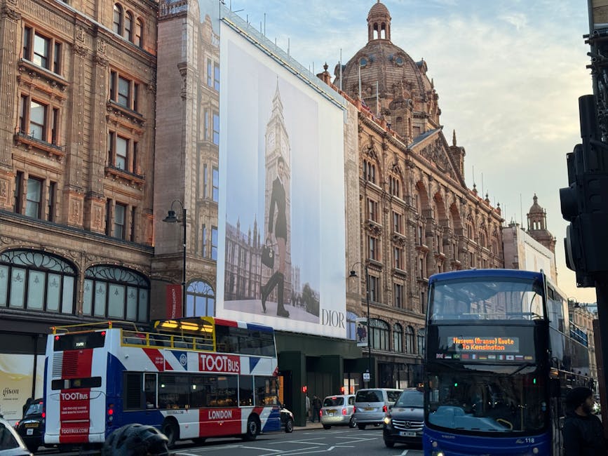 A black and white photograph of a historic urban building with ornate architectural details, including decorative cornices, tall windows, and a rounded corner tower topped with a spire. The building features illuminated signage for a hotel, with the word 'Harrods' vertically displayed on a marquee-style sign along the rounded corner, and a large decorative circular sign with lights above the entrance. The ground floor appears to be a commercial space, with storefront windows and awnings. The scene is set on a city street with parked cars and pedestrians visible at the pavement level. The overall atmosphere is based on the building's classic design and the lively street environment, providing context for how private waste collection and rubbish removal services might be necessary for maintaining such historic urban sites, especially when managing trash during renovations or street events. The muted tones emphasize the architectural features and urban setting, aligning with the context of alternative waste handling in busy city environments.