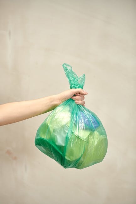 A person's arm is extended holding a green plastic rubbish bag by its tied top. The bag appears partially transparent, with visible contents and a hint of blue items inside. The bag is slightly crumpled, showing its flexible and lightweight plastic material. The background is a plain, neutral-colored wall, with soft, even lighting that highlights the bag's glossy surface. The scene suggests an act of waste disposal or collection, aligning with rubbish removal services, and reflects the practice of private or independent waste handling. The focus is on the bag itself, with no other objects or environmental details visible, emphasizing the straightforward nature of rubbish collection tasks.
