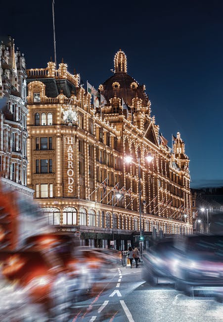 A tall, ornate building illuminated with numerous warm white string lights along its architectural features, such as turrets, balconies, and decorative facades, stands prominently in the scene. The building's exterior showcases a classic, historic style with intricate detailing, large windows, and a rounded central tower topped with a decorative dome. To the left of the building, a vertical sign displaying the name 'HARDRODS' is affixed. In the foreground, blurred red vehicle taillights and dark-colored cars create streaks across the street, indicating motion during nighttime. A couple of pedestrians are visible crossing the road beneath streetlights, which cast a warm glow on the pavement. The sky overhead is a deep, dark blue, providing a contrasting backdrop to the brightly lit building. The scene captures a lively urban environment at night, with the lighting emphasizing the building's elaborate architecture, reminiscent of a historic or luxury retail area involving private or independent collection of waste, aligning with the context of rubbish removal and alternative waste handling in urban settings. The environment appears clean and well-maintained, with the street surface clear of debris, suitable for professional rubbish removal or on-site clearance services as offered by [COMPANY_NAME].