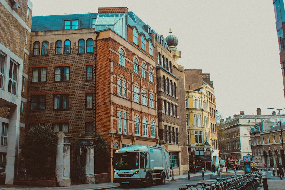 A street scene in an urban area featuring several multi-storey buildings with varied architectural styles. The central building is constructed with reddish-brown brickwork, prominent arched windows with white frames, and a decorative cornice. To its right, there is a cream-colored building with ornate window moldings and a rounded turret topped with a dome-like structure. The street includes a white waste collection truck parked along the curb, partially visible in the foreground, indicating the presence of rubbish removal services. The pavement is lined with black bollards and bicycle racks, with additional vehicles and street lamps in the background. The environment appears well-lit by natural daylight, capturing a typical cityscape that involves alternative waste handling and private disposal activities, exemplified by the parked waste collection vehicle and the tidy appearance of the street. The scene reflects an active, built-up urban setting suitable for independent rubbish removal and waste management solutions, such as those offered by companies operating in London districts like Knightsbridge.