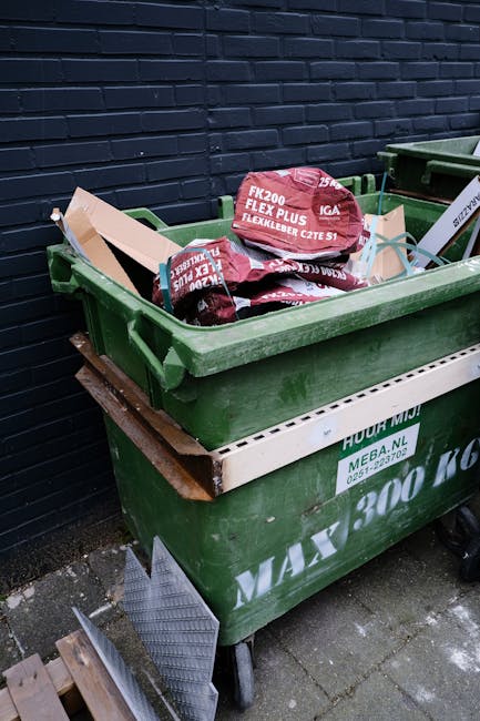 A green wheeled skip filled with red and black bags of garden or construction waste, positioned against a dark blue brick wall on a paved surface. The bags are partially stacked and appear to contain soil, mulch, or similar materials, with some bags showing text indicating their contents. The skip is partially covered with a wooden board and has a white label with green text on the side. Nearby, a metal grating and some small wooden blocks are visible on the ground, suggesting an area designated for bulk waste or garden refuse collection. The scene is outdoors, with natural lighting illuminating the waste materials and the surrounding environment, reflecting a typical private refuse disposal setup. The presence of the skip and waste bags aligns with alternative waste handling methods, supporting independent rubbish removal services such as those offered by companies like rubbishcollectionknightsbridge.org.uk.