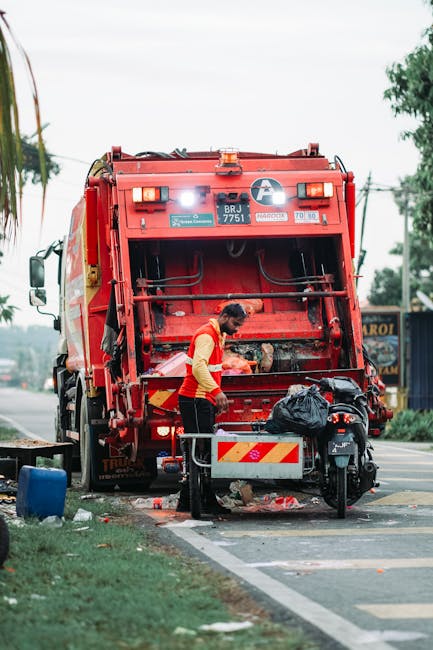 A waste collection worker, dressed in a red and yellow high-visibility jacket, is seen operating a large red rubbish truck parked on the side of the road near a curb. The back of the vehicle is open, revealing mechanical components and a compartment for waste. The worker stands close to a small black motorcycle parked next to the truck, with a black trash bag placed nearby. The scene includes typical roadside surroundings with a grassy verge, a blue trash bin on the ground, and a few scattered pieces of rubbish, indicating an active rubbish removal operation. In the background, there are trees, utility poles, and a building with a sign, suggesting a commercial or suburban area. The lighting appears natural, with an overcast sky, providing neutral and even illumination across the scene. The image exemplifies on-site waste collection, potentially part of an alternative rubbish removal service that bypasses standard council collection methods, emphasizing private waste handling efforts.