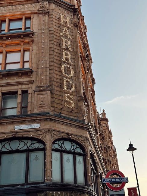 The image depicts a historic multi-storey building with ornate architectural details and a stone façade, featuring large arched windows on the ground floor with decorative metal framing. The building's exterior wall has a vertically mounted sign made of individual illuminated letters spelling 'FEARROADS' attached to the brickwork. The façade includes classical decorative elements such as cornices, pilasters, and intricate stone carvings. To the right of the building, a tall street lamp with a traditional lantern design is visible, along with a London Underground sign indicating proximity to an underground station. The sky above is clear with a few scattered clouds, and natural daylight illuminates the scene, highlighting the building's textured stone surface and the signage. The overall setting suggests an urban environment, possibly in a historic district, with occasional visual references to private or independent businesses, aligning with themes of alternative or private waste management options that might operate within such areas. This detailed scene provides an appropriate context for waste collection and rubbish removal services associated with city environments or buildings of similar architectural style, as seen through [COMPANY_NAME].