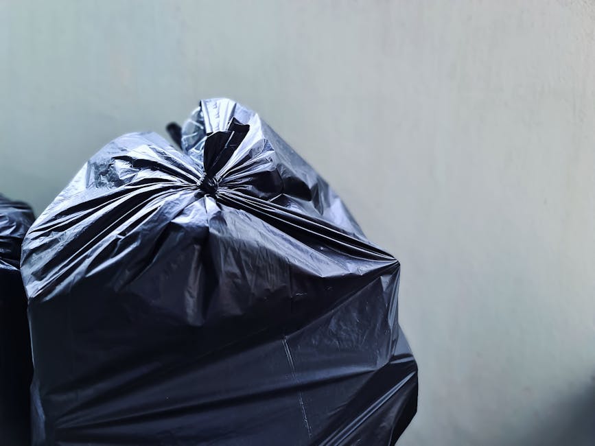 A close-up image showing multiple large black plastic rubbish bags stacked together in an indoor environment. The bags are tightly knotted at the top, with some crinkling and shiny textures visible on their surfaces, indicating they are made of durable garbage film. The background consists of a plain, light-colored wall with minimal detail, suggesting a utilitarian space such as a storage room or a back area where waste is temporarily collected before removal. The bags appear filled to capacity, with a few bottles or other small items slightly visible through the top openings of some bags. The scene reflects typical arrangements for private waste disposal or on-site clearance, often handled by independent waste collection services like those provided by firms such as rubbishcollectionknightsbridge.org.uk, focusing on waste that requires alternative management outside municipal bins.