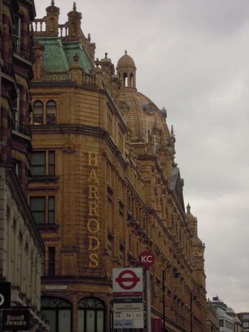 The image features a historic multi-storey building constructed from warm-toned sandstone, with elaborate architectural details including decorative cornices, columns, and a prominent dome at the top. The facade displays numerous windows with dark frames and some have small balconies, adding to the ornate appearance. Vertical signage reading 'HARRANDS' is affixed to the side of the building, indicating a business presence. In the foreground, a red and white bus stop sign is visible, along with part of a neighboring building on the left, which appears to be constructed from similar materials. The sky overhead is overcast with thick grey clouds, creating a subdued atmosphere. The street scene suggests an urban environment, with hints of additional modern structures in the distance, while the overall setting reflects a mix of historic architecture and contemporary city life. The detailed textures of the stonework and the atmospheric lighting emphasize a sense of classical London architecture, aligning with the context of alternative waste management and city infrastructure nearby.